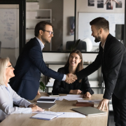 Geschäftstreffen mit Handschlag zwischen zwei Personen, während Kolleginnen am Tisch zustimmend zuschauen.