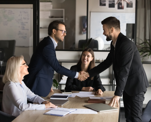 Geschäftstreffen mit Handschlag zwischen zwei Personen, während Kolleginnen am Tisch zustimmend zuschauen.